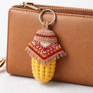 Puede incluir: Un llavero artesanal de ganchillo con forma de seta. El llavero presenta un tallo amarillo y un sombrero estampado en rojo, beige y amarillo. El llavero está sujeto a un anillo metálico y se muestra junto a una cartera de cuero marrón.