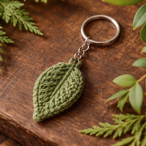 May include: A green crocheted leaf keychain with a silver key ring. The leaf has detailed stitching and a realistic vein pattern. The keychain is set against a wooden background with green foliage.