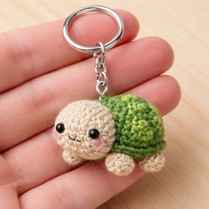 Può includere: Un portachiavi a forma di tartaruga all'uncinetto. La tartaruga ha testa e arti beige, guscio verde e occhi neri con guance rosa. È attaccato a un portachiavi e una catena color argento. Il portachiavi è tenuto in mano.
