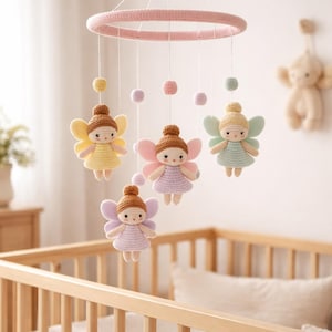 May include: A handmade crochet baby mobile featuring four fairy dolls with colorful wings and dresses. The mobile has a pink ring and is suspended above a wooden crib. The fairies are in yellow, pink, and green.