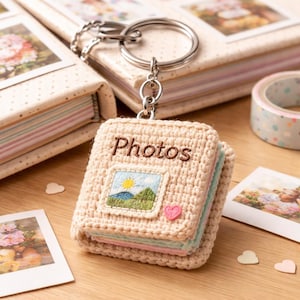 Puede incluir: Un mini llavero de álbum de fotos de ganchillo beige con la palabra "Photos" bordada en la portada. La portada presenta una pequeña escena de paisaje bordada y un corazón rosa. El llavero está unido a un anillo de metal plateado.
