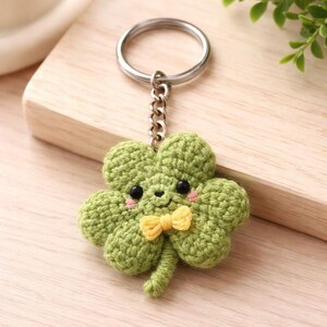 May include: A green crocheted four-leaf clover keychain with a smiling face, black eyes, pink cheeks, and a yellow bow tie. The keychain has a silver metal ring and chain. The clover is made of a textured yarn.