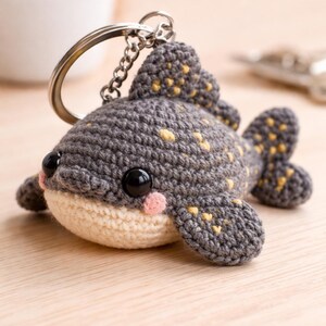 May include: A handmade crochet keychain in the shape of a fish. The fish is gray with yellow spots and black eyes. It has a cream-colored belly and pink cheeks. The keychain is attached to a silver metal ring and chain.