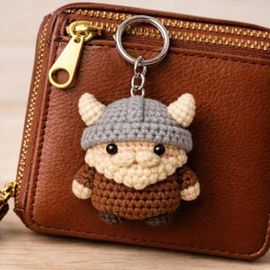 May include: A handmade crochet keychain featuring a Viking character with a gray helmet, beige beard, and brown tunic. The keychain is attached to a silver ring and rests on a brown leather wallet with a gold zipper.