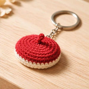 May include: A handmade red and white crocheted keychain. The keychain features a circular design with a red top and a white bottom. The keychain is attached to a silver metal ring and chain.