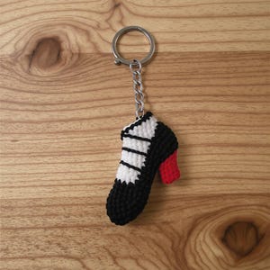 以下が含まれることがあります： 手作りのハイヒール型の編み物キーホルダー。靴は黒を基調とし、白のアクセントと赤いヒールが特徴です。キーホルダーはシルバーの金属リングとチェーンに取り付けられています。