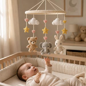 May include: A crochet baby mobile featuring a circular frame with hanging elements including white clouds, yellow stars, pink hearts, and three stuffed animals: a light brown bear, a grey elephant, and a cream-coloured rabbit with long ears.