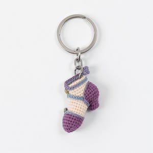 以下が含まれることがあります： 手作りのキーホルダー。かぎ針編みのミニチュアハイヒールシューズが特徴です。靴は主にクリーム色で、ヒールとつま先を含む紫色のアクセントがあります。ライトブルーとゴールドのディテールがデザインを補完しています。キーホルダーリングはシルバーです。