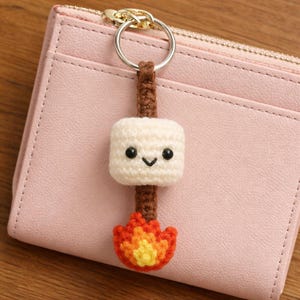 Puede incluir: Un llavero de ganchillo hecho a mano con un malvavisco sonriente en un palo sobre una fogata. El malvavisco es blanco con ojos negros y una sonrisa cosida. Las llamas son naranjas y amarillas. El llavero está sujeto a un anillo plateado.