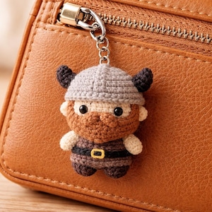 May include: A handmade crochet keychain featuring a Viking figure. The Viking has a gray helmet with horns, a brown beard, and a belt with a gold buckle. The keychain is attached to a silver metal ring. The background is a brown leather wallet.