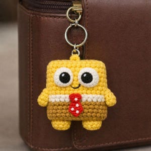 Puede incluir: Un llavero amarillo de ganchillo con forma de personaje de dibujos animados, con grandes ojos con borde blanco y una corbata roja con lunares. El llavero está sujeto a un anillo de metal, sobre un fondo de cuero marrón.