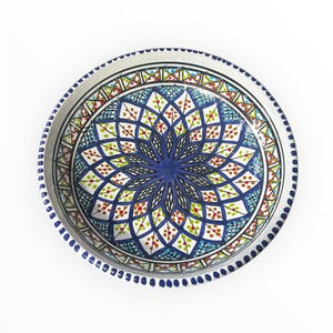 May include: A round, hand-painted ceramic plate with a vibrant floral design. The plate features a central blue flower surrounded by intricate patterns in blue, green, red, and white. The rim is decorated with a dotted border.