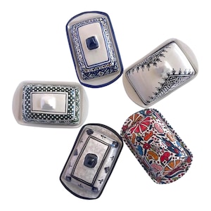 May include: A selection of ceramic butter dishes with lids. The dishes display diverse patterns, such as floral, geometric, and abstract designs. Colours include blue, green, white, black, red, and orange. Each dish has a rectangular shape with a lid and a handle.