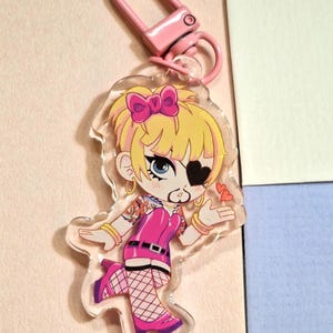 Könnte beinhalten: Ein klarer Acryl-Schlüsselanhänger mit einer Cartoonfigur mit blonden Haaren, einer rosa Schleife und einer schwarzen Augenklappe. Die Figur trägt ein rosa Outfit mit Netzstrümpfen und rosa Stiefeln. Oben ist ein rosa Verschluss befestigt.