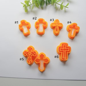 May include: Six orange plastic cross-shaped cookie cutters, numbered 1 through 6, are arranged on a white surface. The cutters vary in design, with some featuring detailed cutouts. Green sprigs are in the background.