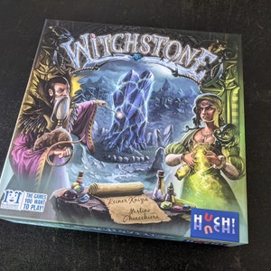 May include: A board game box with a fantasy theme. The box features a blue crystal, a wizard with a white beard, and a woman holding a potion. The game is called "Witchstone" and is made by "Hoch! Chi".