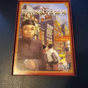 May include: A red box with a gold border and the words "Chinatown: The Art of Trading" on the front. The box features an illustration of a man in a traditional Chinese outfit standing in front of a cityscape.