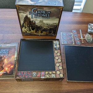 May include: A Game of Thrones board game box set with a black game board, a black player board, and a variety of game pieces. The box art features a castle and the text "A Game of Thrones The Board Game".