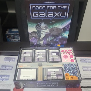 May include: A board game titled "Race for the Galaxy" with a box featuring a space scene. The open game box reveals cards, tokens, and dice. Additional game components and instructions are visible on a red surface.