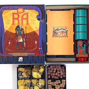 May include: Opened board game box titled "RA: Pharaoh Edition". The box art depicts an Egyptian theme with the word "RA" prominently displayed. Inside are teal tokens, wooden blocks, and gold game pieces. The game is set in a box.
