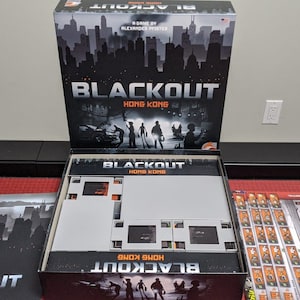 May include: An open box of the board game Blackout: Hong Kong. The box is black with a cityscape silhouette and the game title in white. The game board is visible inside the box, along with various game components.