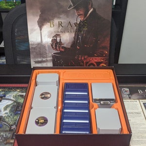 May include: An open box with a game board and game components for the board game Brass: Lancashire. The box has a blue and orange insert with compartments for the game pieces. The game board is a map of Lancashire, England. The game components include cards, tokens, and dice.