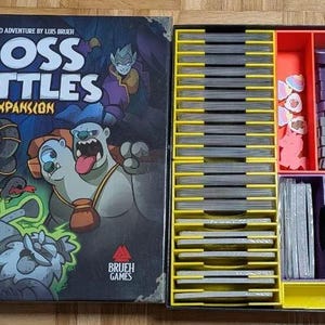 May include: The open box of the Boss Battles Expansion board game. The box art features cartoon monsters and the game title. The inside of the box has compartments with game pieces and cards. The compartments are yellow, red, and purple.