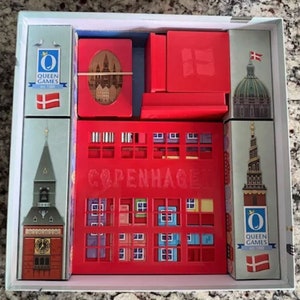 May include: A red and white board game box with the title "Copenhagen" and a picture of a city skyline. The box includes a variety of colourful tiles and cards. The game is made by Queen Games.