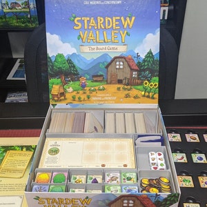 May include: An open box containing a Stardew Valley board game insert. The insert has compartments for cards, dice, and other game components. The box lid features a colourful illustration of the game's setting.
