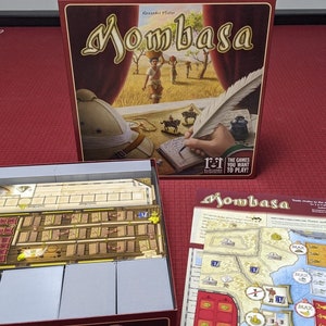 May include: The board game "Mombasa" is shown, with a box displaying artwork of figures in a field and the game's title. Game components, including a board and cards, are visible. The box is on a red surface.