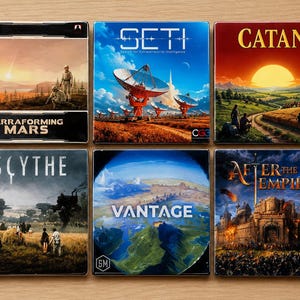 May include: Six board game boxes are displayed, including "Terraforming Mars," "SETI," and "Catan." The boxes feature colorful artwork depicting landscapes, futuristic scenes, and historical settings. The games are arranged on a light wood surface.
