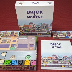 May include: A board game titled "Brick & Mortar" with a box, game board, cards, and rule book. The box and rule book feature a colorful illustration of a street with shops and houses. The game board has compartments for game pieces.