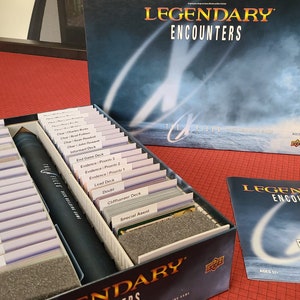 May include: A close-up of a game organizer for the board game "Legendary Encounters: An Alien Deck Building Game". The organizer is made of cardboard and has slots for all of the game's cards, including the character decks, the encounter decks, the special asset decks, and the villain decks. The organizer also has a slot for the rule book. The organizer is sitting on a red table.