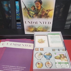 May include: A board game called "Undaunted: Reinforcements" with a military theme. The box shows soldiers and a tank. Inside the box are cards, tokens, and rule books. The game components are neatly arranged in a pink box.