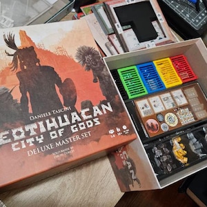 Puede incluir: Caja de juego de mesa abierta titulada "Teotihuacan: City of Gods Deluxe Master Set". La caja es naranja y marrón con una ilustración de guerrero. En el interior hay piezas de juego, incluidos divisores de plástico de colores y figuras en miniatura.