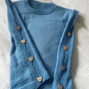 Pode incluir: Um suéter de tricô azul claro com botões em forma de urso nas mangas.