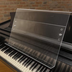 May include: A clear acrylic music stand extension resting on a black grand piano. The rectangular transparent panel features a horizontal shelf at the base and is secured with visible metal hardware, including screws and circular fasteners on the sides.