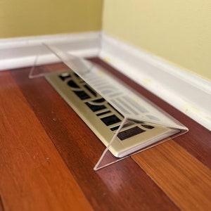 May include: A clear plastic vent cover protects a floor vent from debris. The vent cover is triangular in shape and sits on a wooden floor.