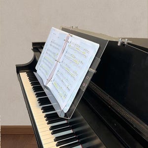 Könnte beinhalten: Ein schwarzes Klavier mit weißen und schwarzen Tasten. Noten werden auf einem transparenten Notenständer aus Acryl angezeigt. Der Notenständer ist am Klavier befestigt. Die Noten sind geöffnet und sichtbar.