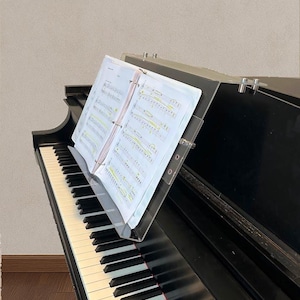 May include: A black piano with white and black keys. Sheet music is displayed on a clear acrylic music stand. The music stand is attached to the piano. The sheet music is open and visible.