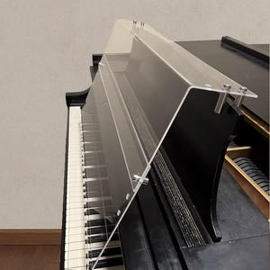 Könnte beinhalten: Eine transparente Klaviertastaturabdeckung aus Acryl, nach rechts abgewinkelt und mit silberner Hardware befestigt. Die Abdeckung befindet sich über den schwarzen und weißen Tasten eines schwarzen Klaviers. Das Klavier steht vor einer neutralen Wand.
