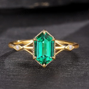 Puede incluir: Un anillo de oro con una gran piedra preciosa verde esmeralda y dos diamantes más pequeños a los lados. El anillo está sobre un fondo negro.