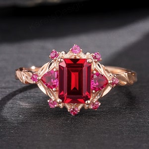 Peut inclure: Une bague en or rose ornée d'une grande pierre rouge foncé rectangulaire au centre, flanquée de pierres roses et rouges plus petites. La bague a une bande détaillée d'inspiration florale et le texte "KARATSDESIGN".