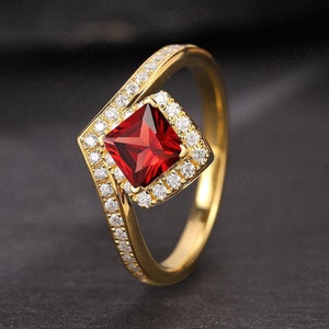 Puede incluir: Un anillo de oro con una piedra roja cuadrada rodeada de piedras transparentes más pequeñas. El anillo tiene un diseño único con una banda curva.