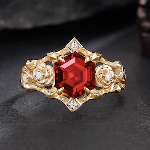 Puede incluir: Un anillo de oro con una gran gema roja facetada en el centro. El anillo tiene un diseño floral con pequeños diamantes y está sobre un fondo oscuro. Los intrincados detalles y el color vibrante del anillo lo convierten en una joya llamativa.