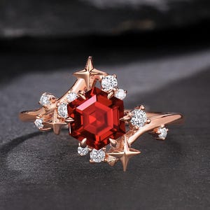 May include: A rose gold ring featuring a large, faceted, hexagonal red gemstone as the centerpiece. The ring is adorned with small, round, clear gemstones and star-shaped accents. The ring is set against a dark background.