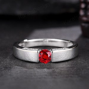 May include: A silver band with a red gemstone and small clear gemstones on the sides. The ring is on a dark gray surface.
