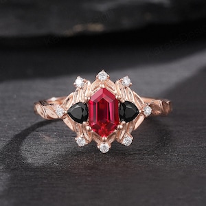 Op de afbeelding: Een verlovingsring van roségoud met een rode edelsteen omringd door kleinere witte edelstenen en twee zwarte edelstenen. De ring heeft een delicaat bloemmotief.