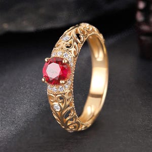 May include: A gold ring featuring a round, faceted red gemstone at the centre, surrounded by small clear stones. The band displays an intricate, openwork scroll pattern along the sides. The text KARATSDESIGN is visible at the top of the image.