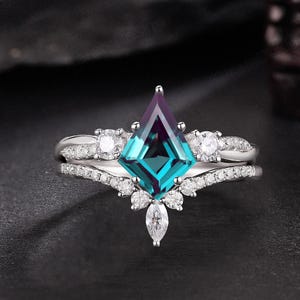 May include: A silver engagement ring set with a teal and purple gemstone in a kite shape, surrounded by smaller white gemstones. The band is also adorned with small white gemstones. The brand name "KARATSDESIGN" is at the top.